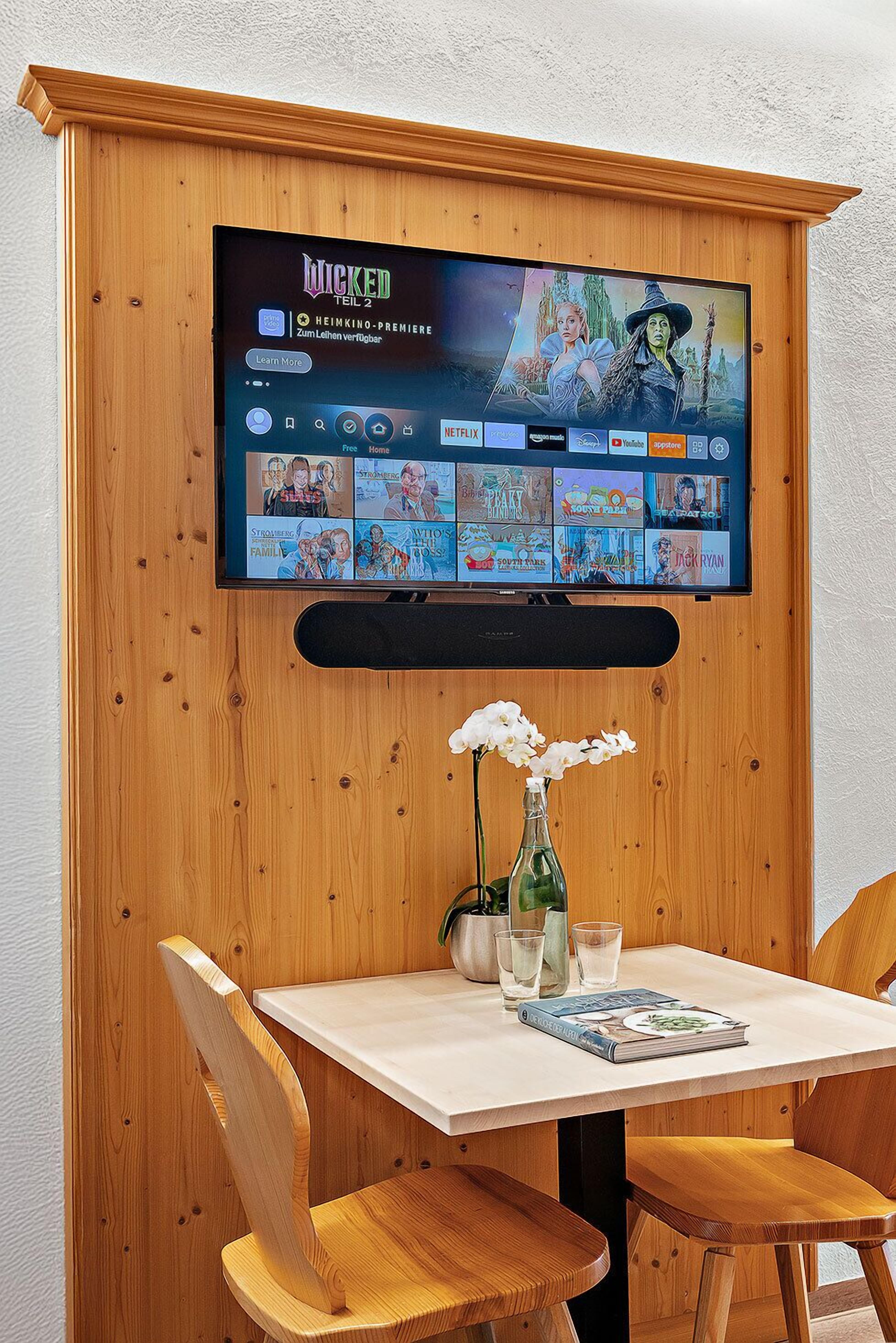Pared de madera con paneles donde se ha montado un televisor que muestra una selección de películas. Debajo del televisor, una mesa con sillas y un jarrón de flores.
