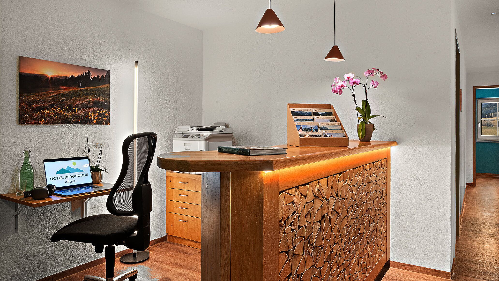 A hotel reception desk with a computer, a chair, a desk lamp, a framed picture, and a plant.