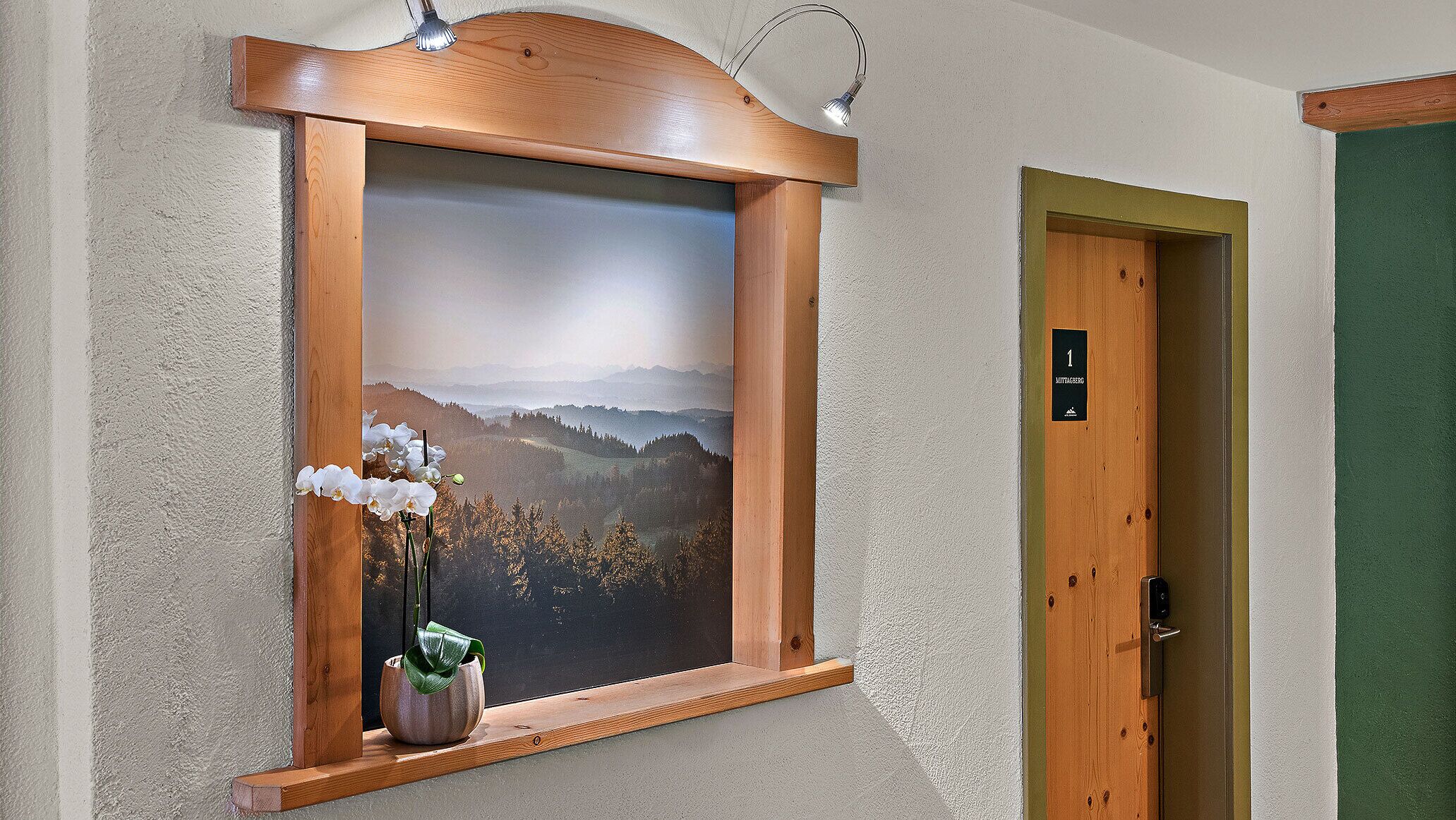 A framed landscape painting with a wooden shelf holding white flowers, a door with a number sign, and a light fixture above the shelf.