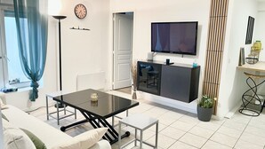 Ein modernes Wohnzimmer mit einer Couch, einem Esstisch mit Glasplatte, einem an der Wand montierten Fernseher und einer Küchenzeile mit Arbeitsfläche und Schränken.