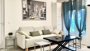 Ein modernes Wohnzimmer mit einer Couch, einem Couchtisch und einem Beistelltisch. An der Wand hängt ein gerahmtes Bild und es gibt ein Fenster mit Vorhängen.
