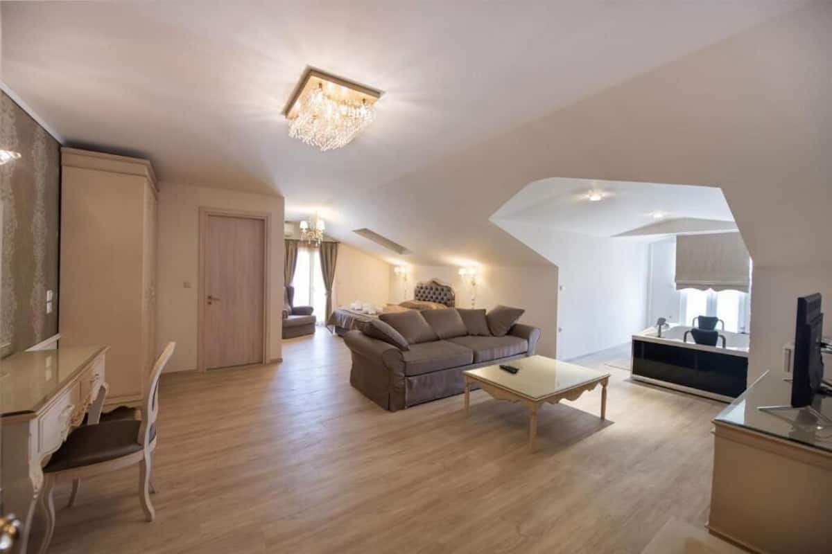 A spacious living room with a sofa, a coffee table, and a TV. There is a dining area with a table and chairs. The room has wooden flooring and a chandelier.