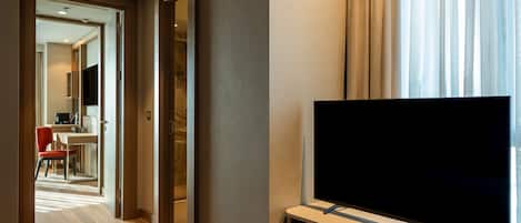 A hotel room with a flat-screen TV on a stand, a wooden floor, and a view of a dining area through an open door.