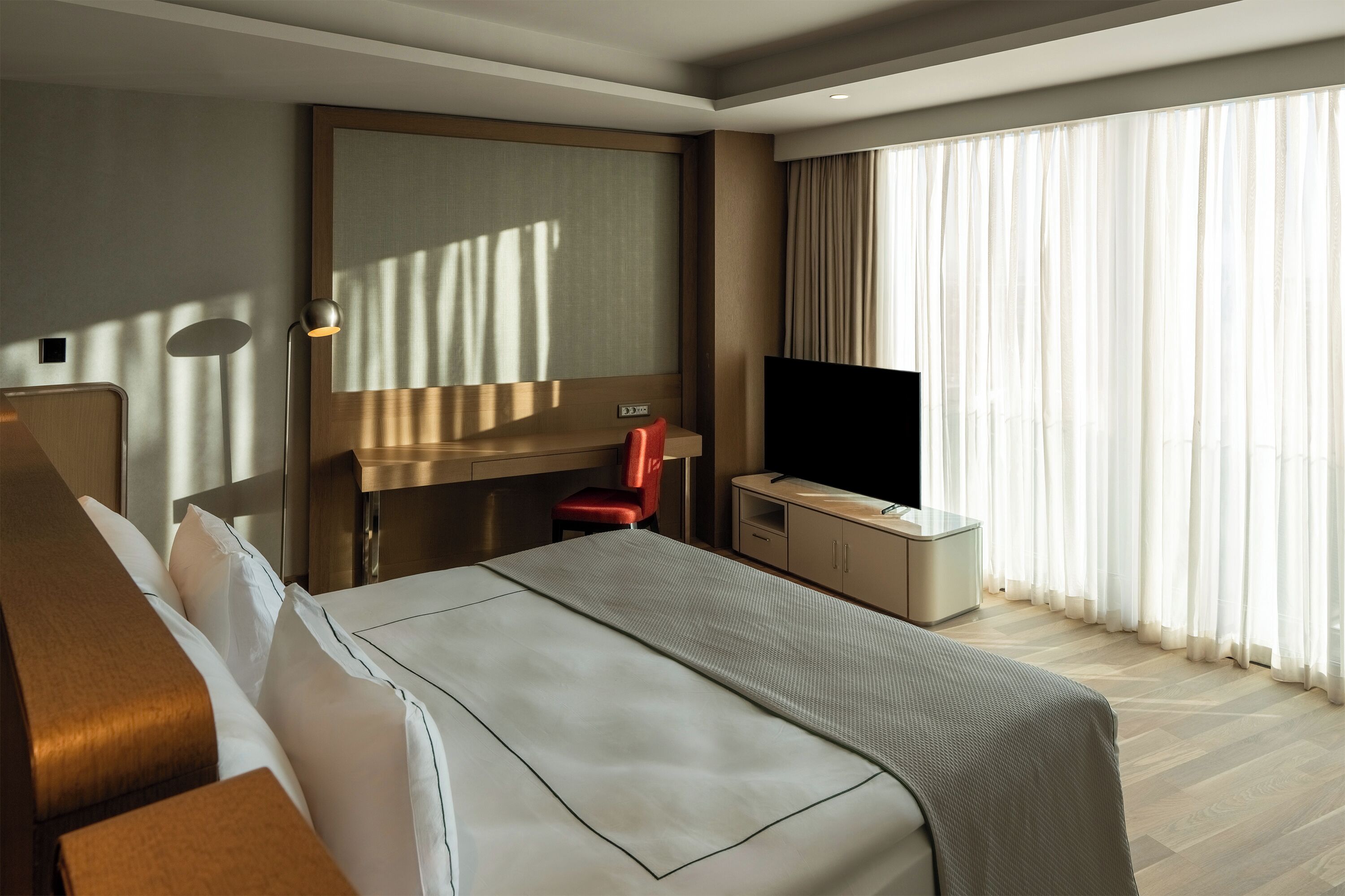 Habitación de hotel con cama, escritorio, silla, televisor y un amplio ventanal con cortinas.