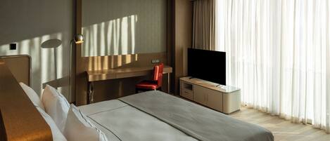 Habitación de hotel con cama, escritorio, silla, televisor y un amplio ventanal con cortinas.