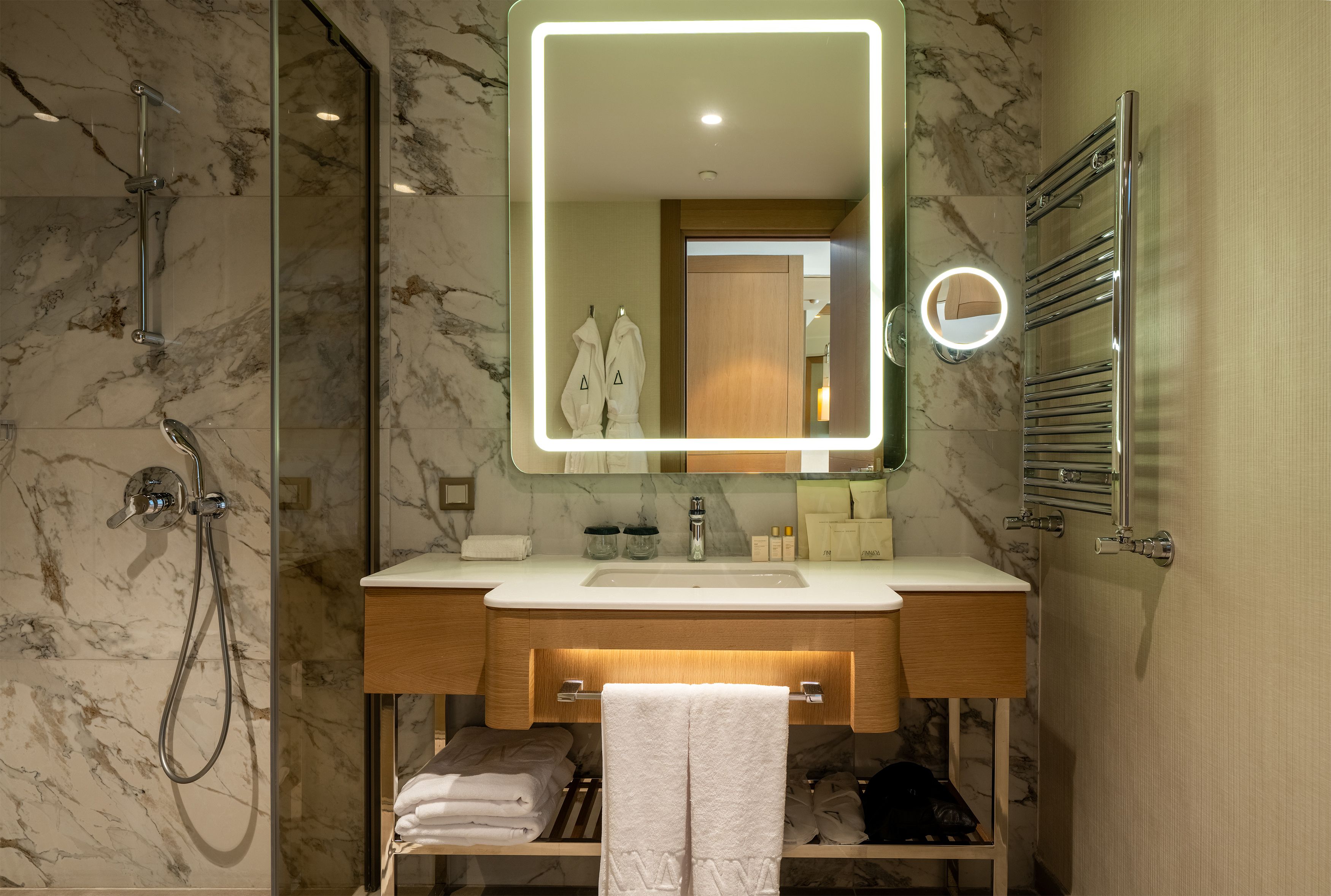 A modern bathroom with a glass-enclosed shower, a marble-tiled wall, a wooden vanity, and a large mirror with a lighted frame.