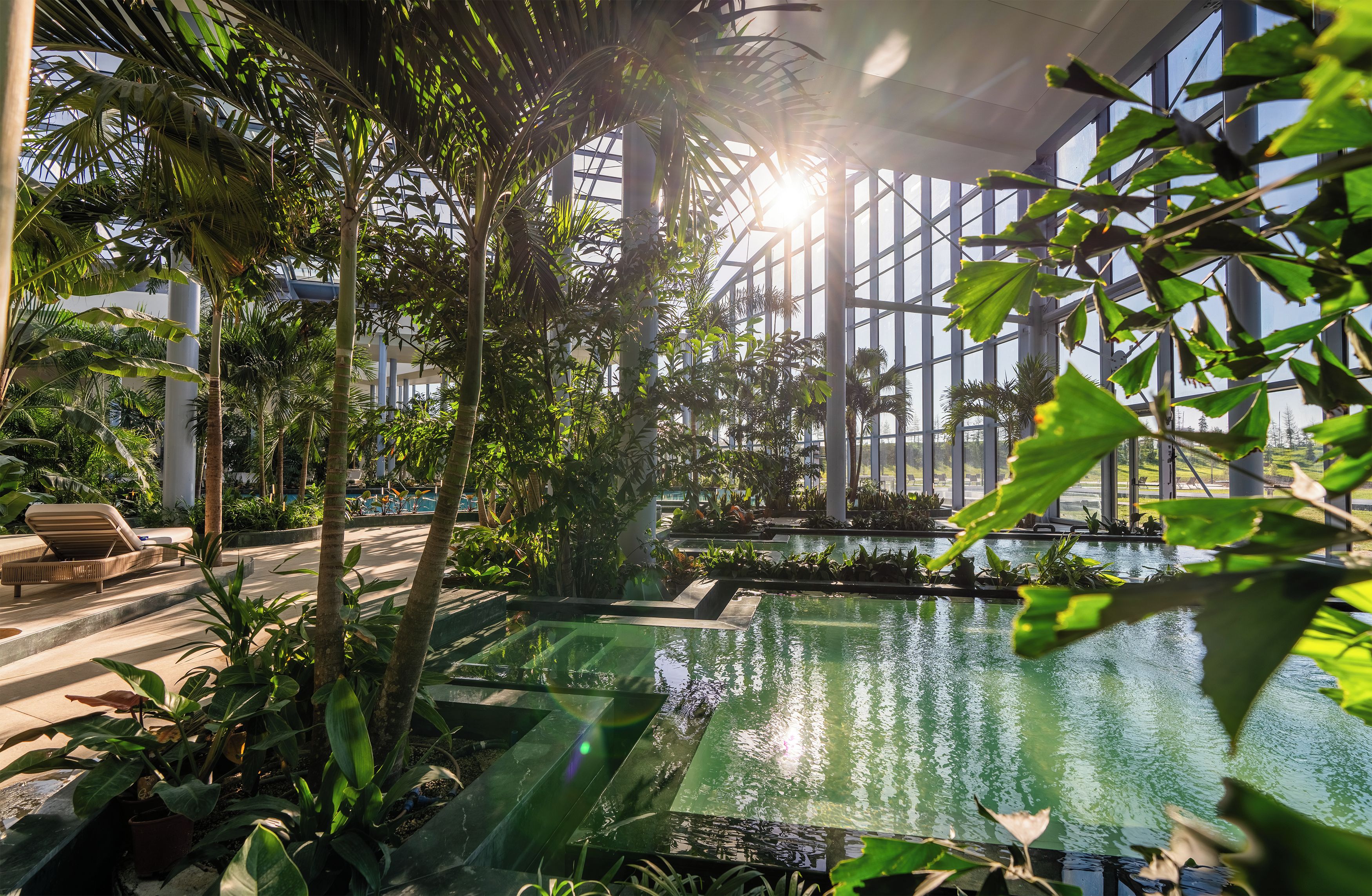 Un jardín interior con fuente de agua, zonas de estar y techo de cristal que deja pasar la luz natural.