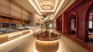 A modern hotel lobby with a central island featuring food and drink stations, a large potted plant, and a red architectural feature on the right. - Sinnada Resort & Thermaland (Afyonkarahisar)