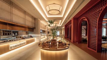 Un moderno vestíbulo de hotel con una isla central que ofrece estaciones de comida y bebidas, una planta grande en maceta y un elemento arquitectónico rojo a la derecha.