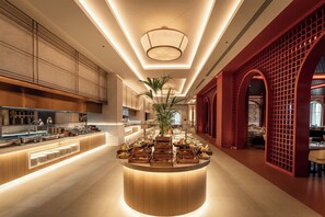 Un hall d’hôtel moderne avec un îlot central comprenant des présentoirs de nourriture et de boissons, une grande plante en pot et un élément architectural rouge situé à droite.