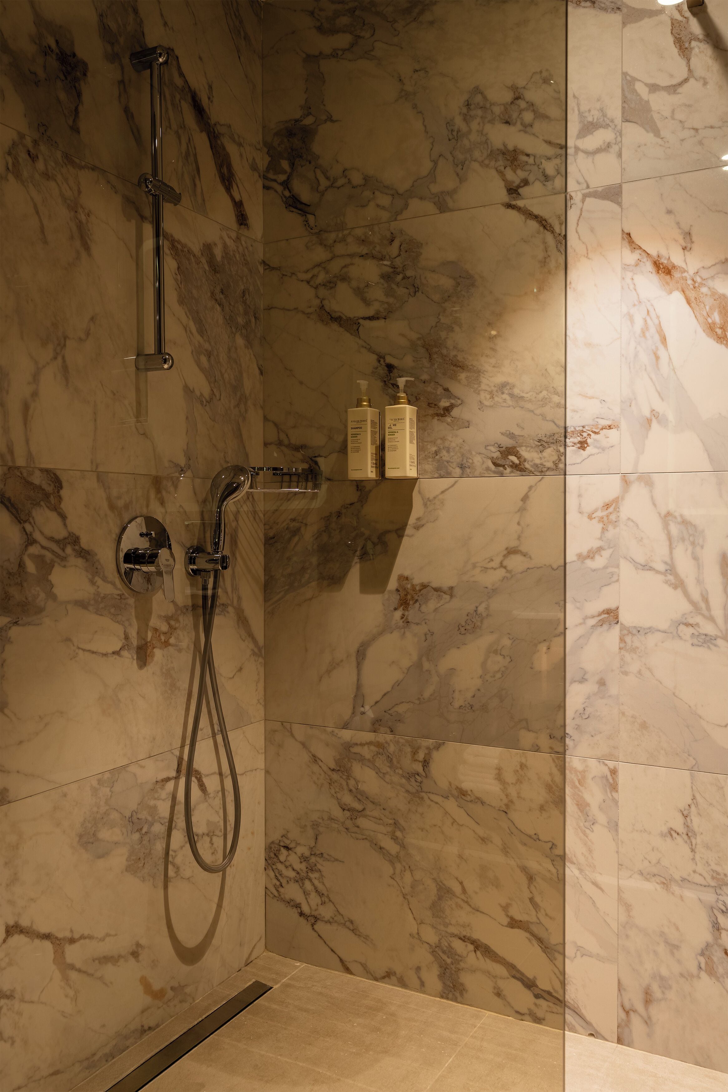 Un baño con ducha, paredes de mármol y un estante con artículos de aseo.