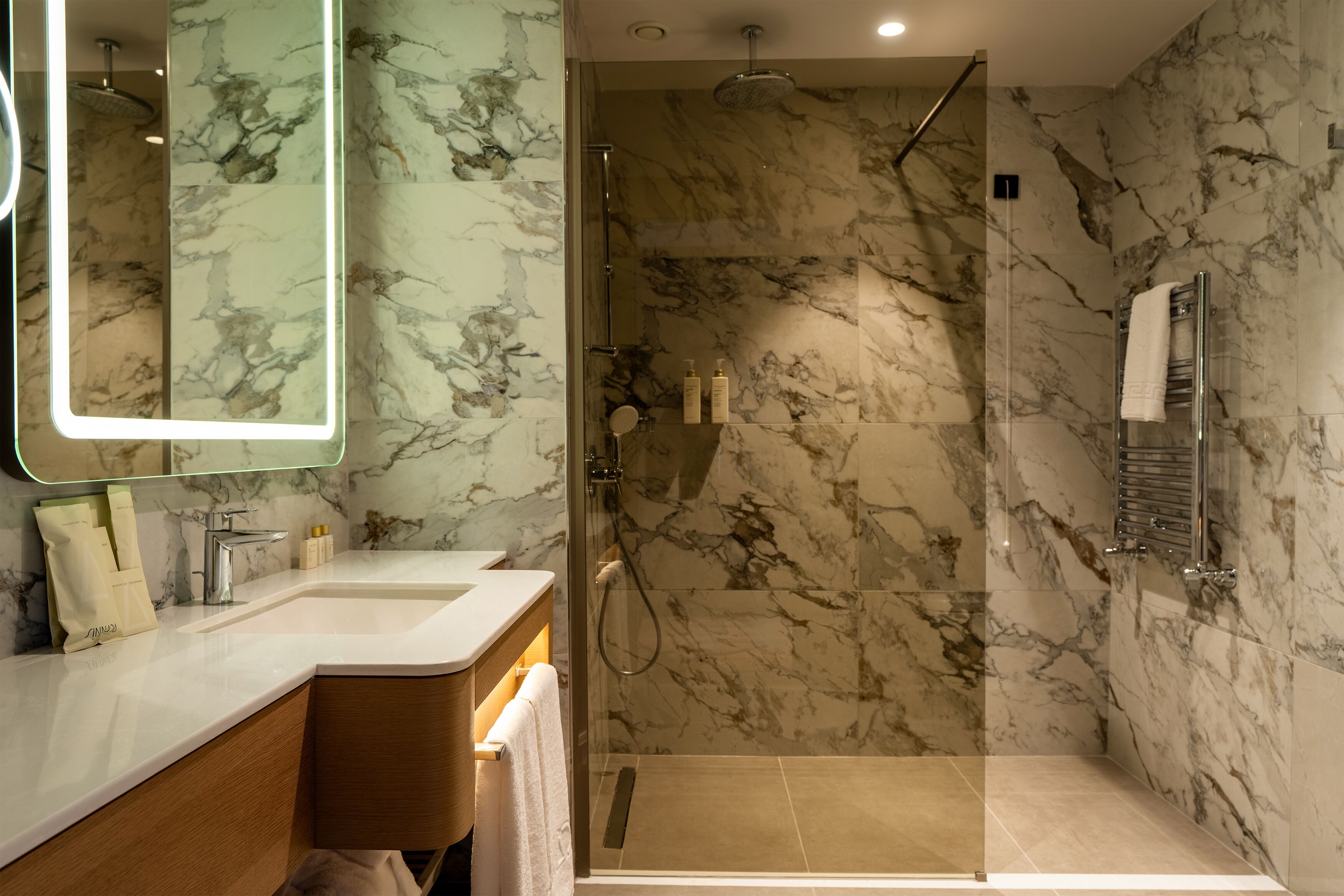 A modern bathroom with a marble shower, a large mirror, and a vanity with toiletries.