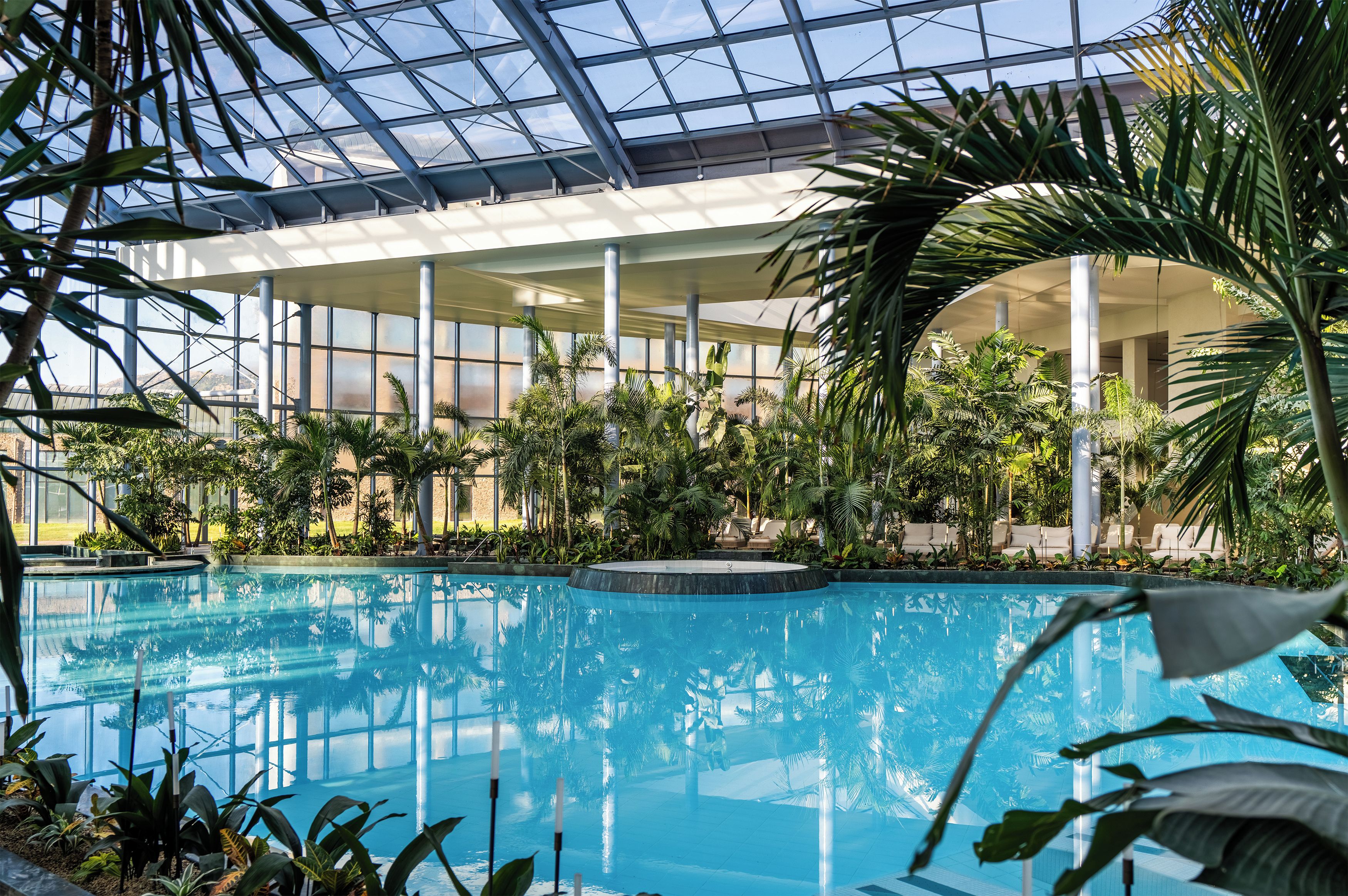 Una piscina interior con techo de vidrio, rodeada de plantas tropicales y zonas de estar.