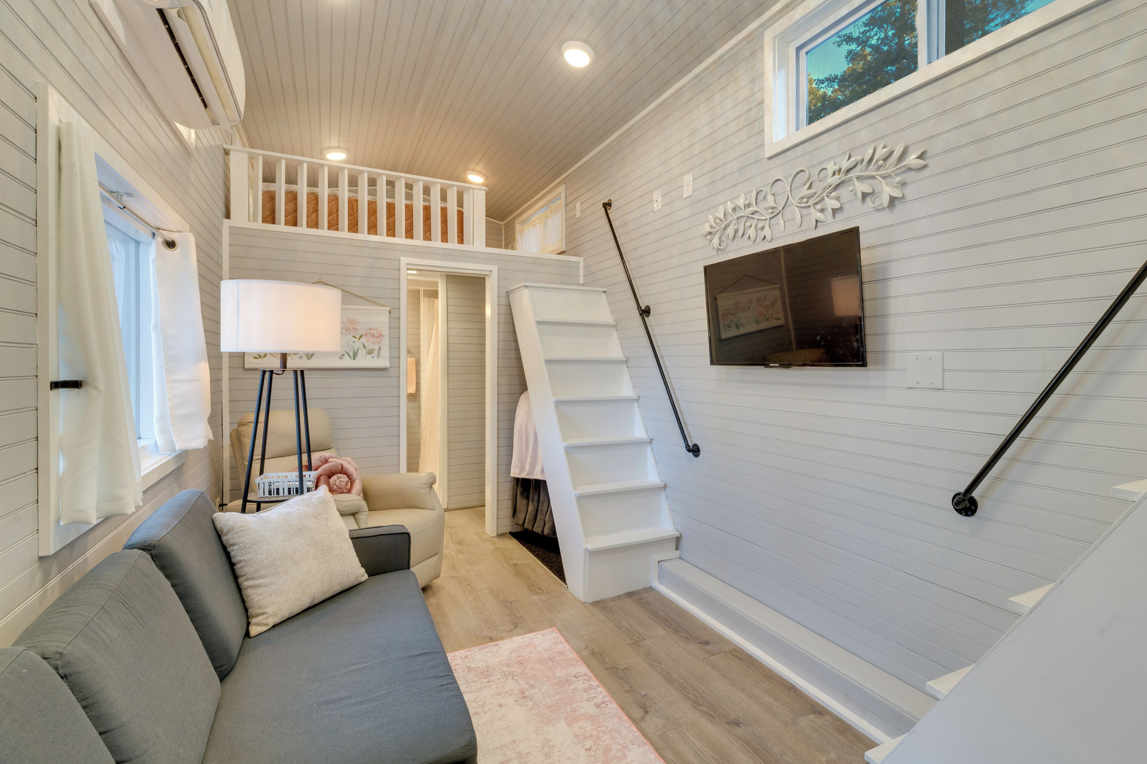 A cozy living room with a gray sofa, a white staircase, and a flat-screen TV mounted on the wall.
