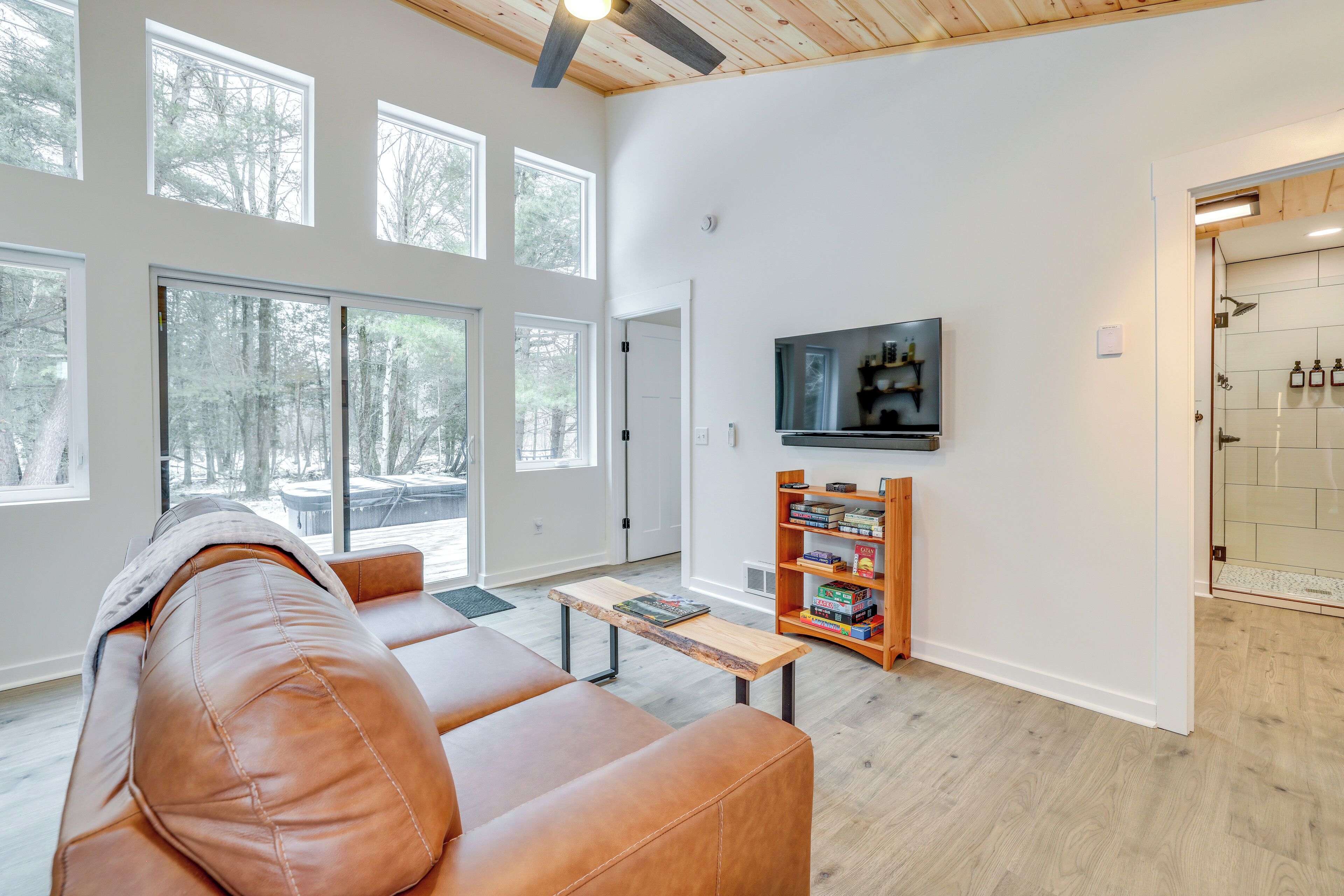 A spacious living room with a leather couch, a wooden coffee table, and a bookshelf. Large windows provide natural light and a view of the outdoors.