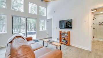 A spacious living room with a leather couch, a wooden coffee table, and a bookshelf. Large windows provide natural light and a view of the outdoors.