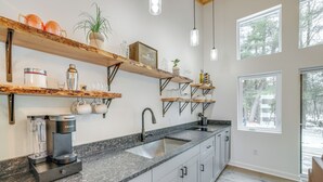 Een moderne keuken met granieten werkbladen, een inbouwspoelbak en roestvrijstalen apparatuur. Daarboven bevinden zich houten planken met diverse keukengerei en een potplant.
