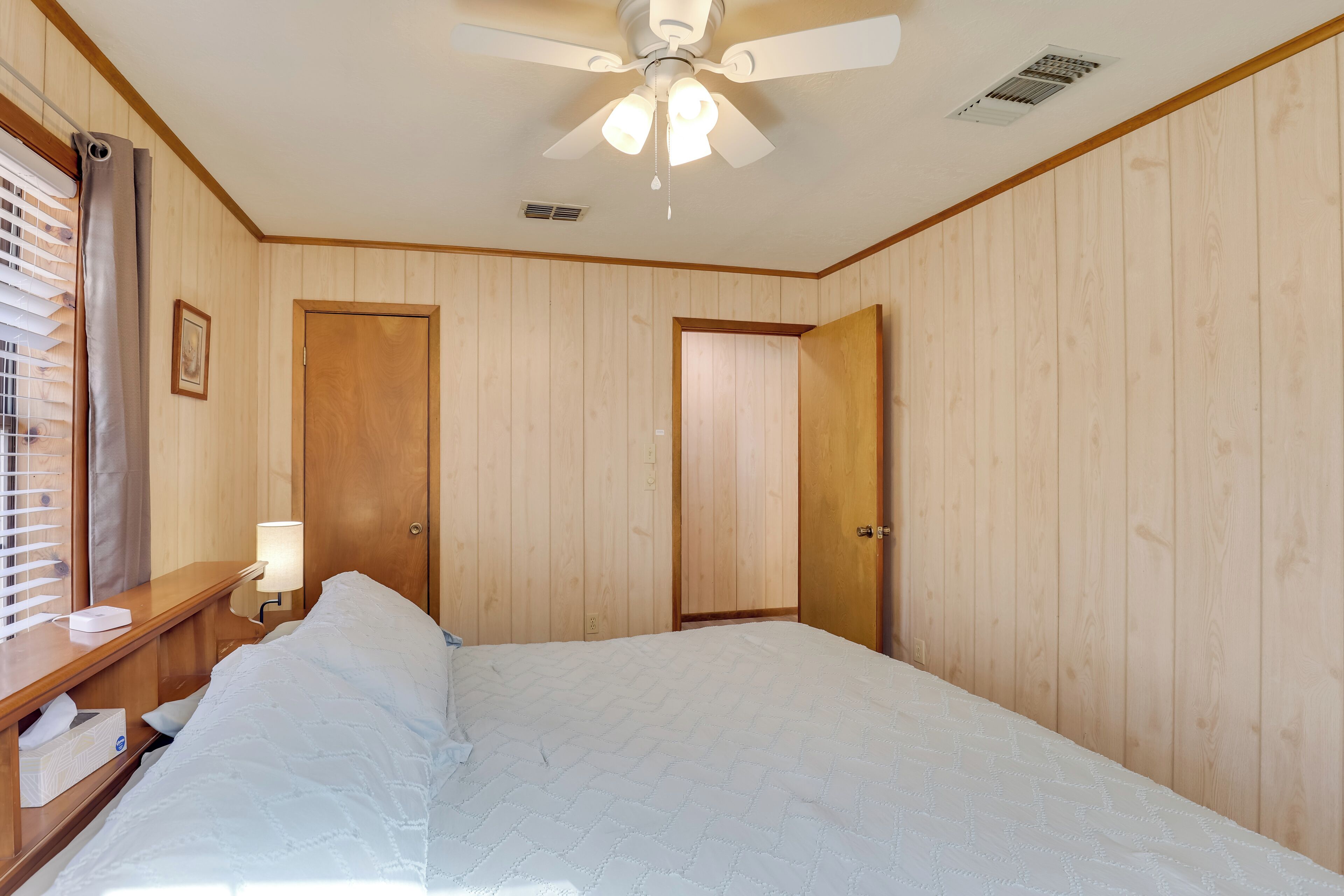 A bedroom with a bed, wooden walls, a ceiling fan, a nightstand, a lamp, and a window with blinds.