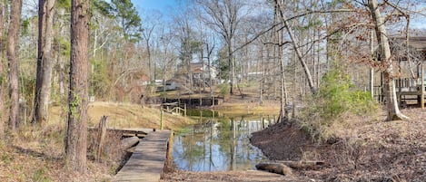 Un pont en bois enjambant un étang, entouré d’arbres et avec un bâtiment visible au loin.