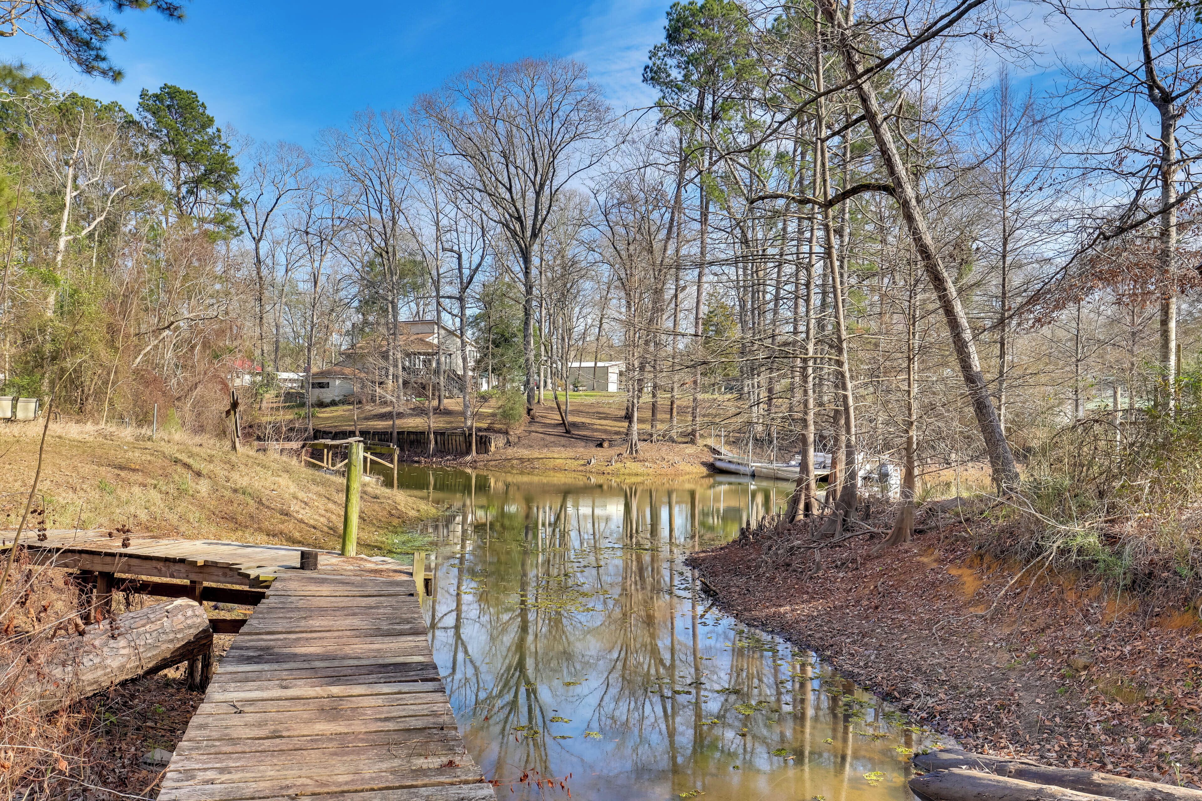A wooden boardwalk leading to a calm pond surrounded by trees and a house in the background.