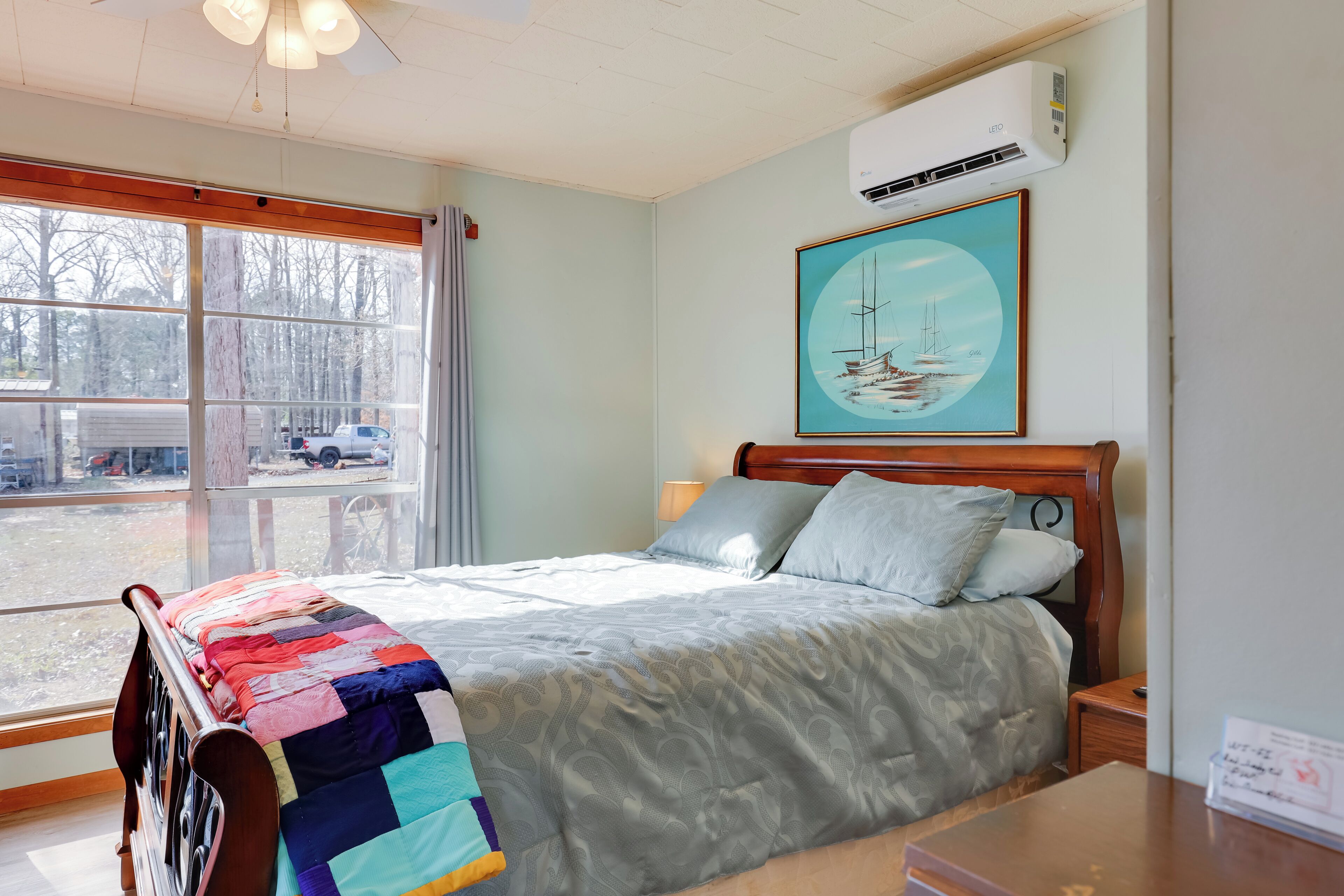 Un dormitorio con un ventanal grande, una cama con colcha colorida, cabecera de madera, mesita de noche y aire acondicionado instalado en la pared.