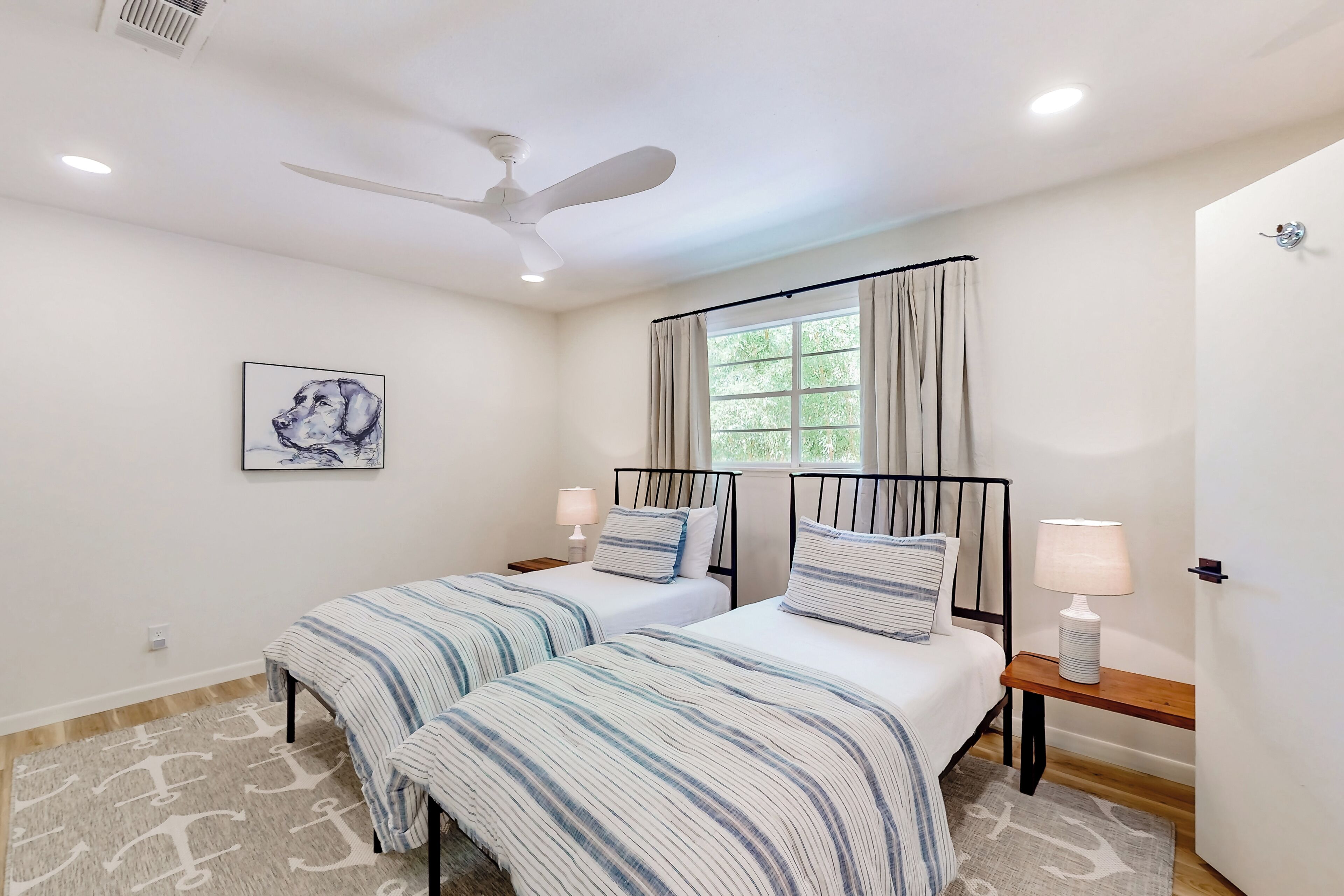 A bedroom with two beds, a ceiling fan, a window with curtains, a painting on the wall, and a rug.