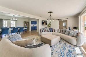 Uma sala de estar espaçosa com um amplo sofá modular, uma área de refeições com cadeiras azuis e uma cozinha ao fundo.