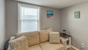 A living room with a beige sofa, a small table with a lamp, and a painting on the wall. - Walk to Public Beach Access! Breezy Hampton Condo (Hampton)