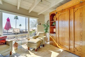 A sunlit living room with a large wooden cabinet, a beige armchair, a glass coffee table, and a view of palm trees through large windows. - Catalina Foothills Gem 8 Mi to Sabino Canyon (Tucson)