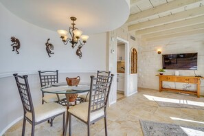 Uma área de refeições com mesa de vidro, quatro cadeiras, um candelabro, obras de arte nas paredes e um console de madeira com uma televisão.