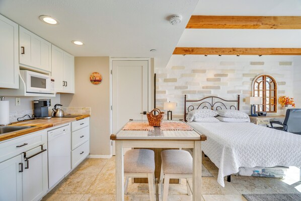A compact kitchen with white cabinets, a microwave, and a coffee maker. There's a dining table with two chairs and a small rug. The room has a bed with white bedding and a nightstand with a lamp. - Hike & Explore Dtwn Tucson! Relaxing Solo Escape (Tucson)