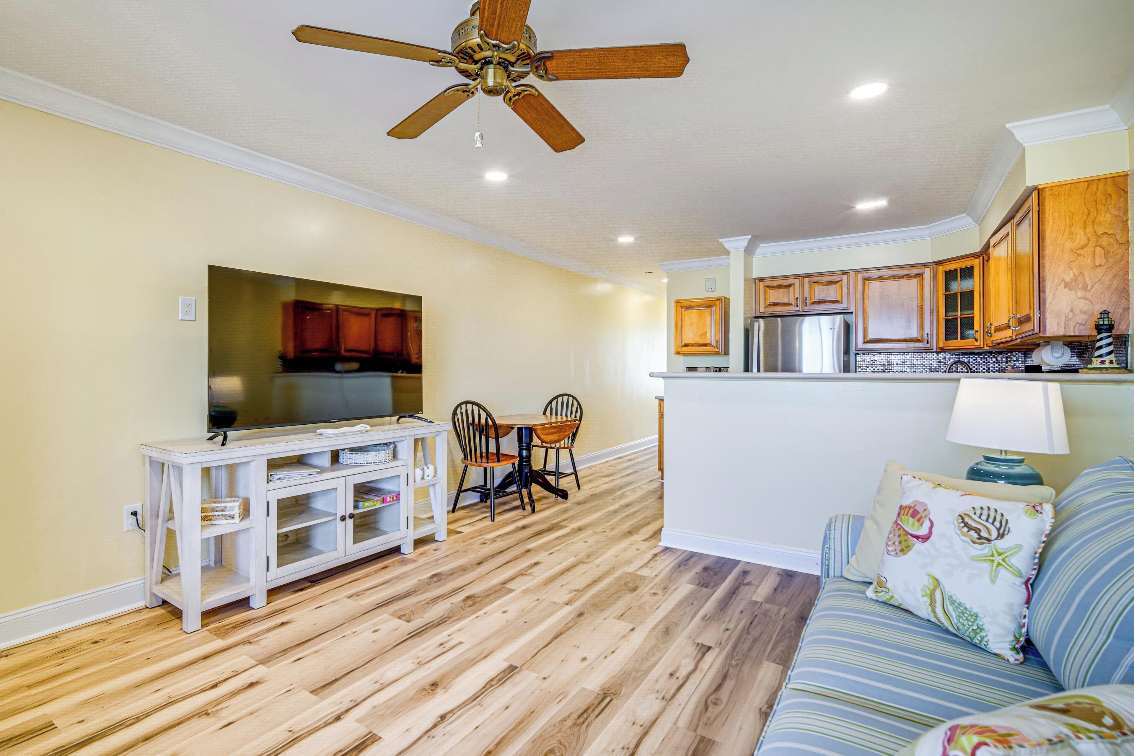 A spacious living area with a wooden floor, a ceiling fan, a dining table with chairs, a sofa with patterned cushions, and a kitchen with wooden cabinets and a built-in refrigerator.