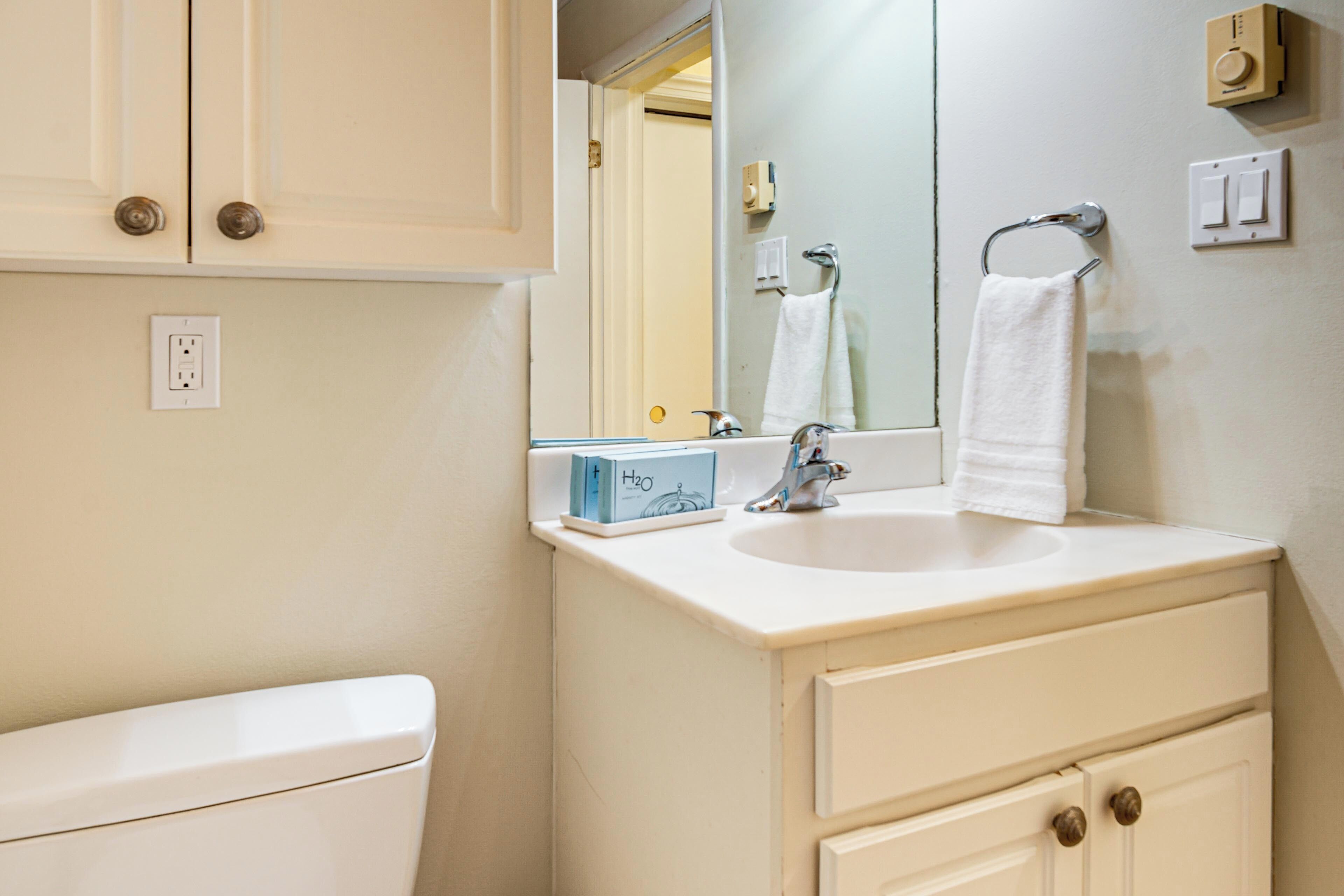 Une salle de bain avec un lavabo blanc, un miroir et un meuble. Une serviette est accrochée au mur et une boîte d’eau se trouve sur le comptoir.