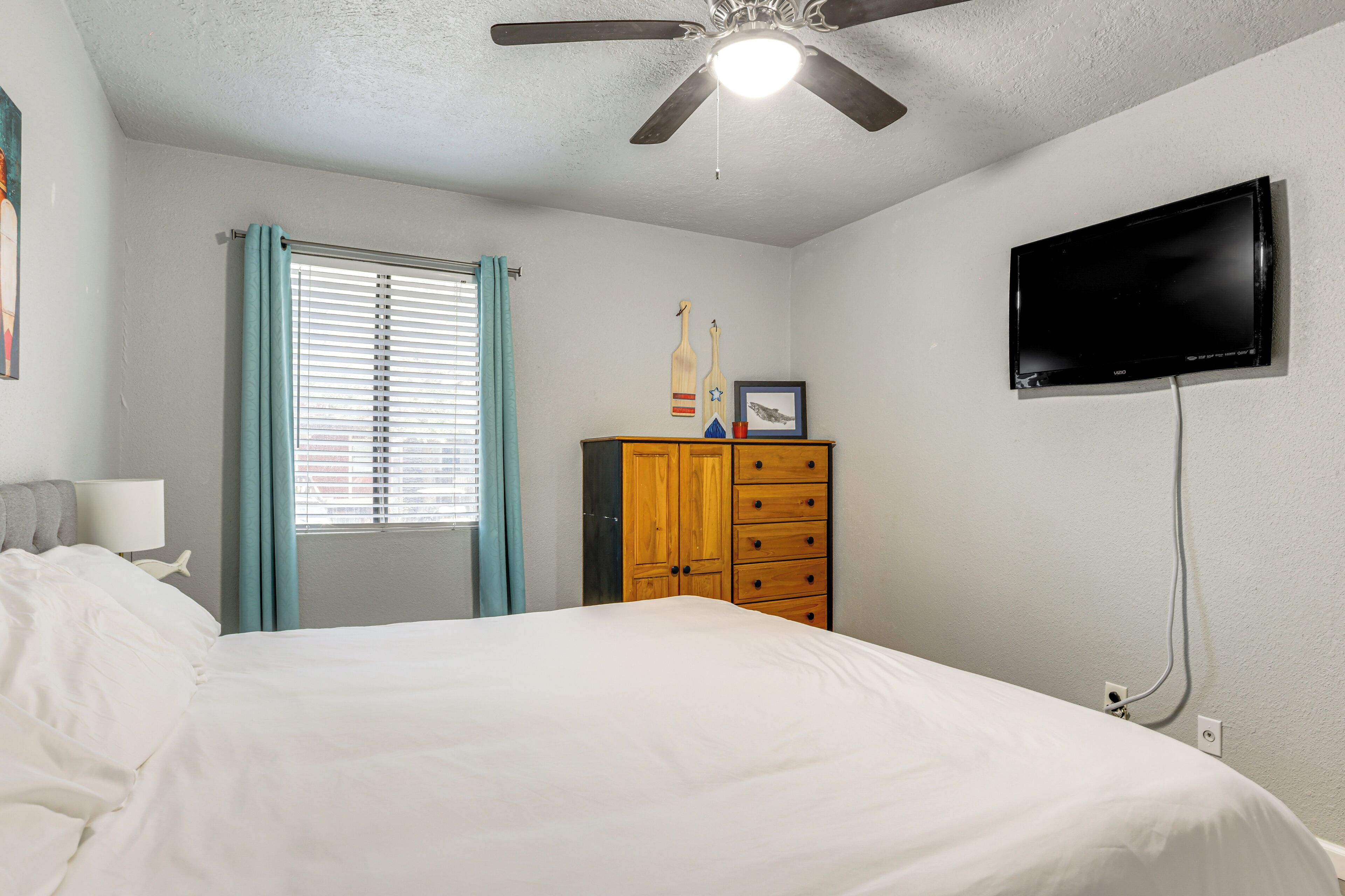 Une chambre à coucher comprenant un lit, une commode en bois, un ventilateur de plafond, une fenêtre avec des rideaux et un téléviseur fixé au mur.