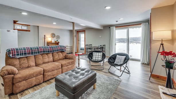 A modern living room with a brown sofa, a black ottoman, a dining table with chairs, and a large window with a view of a snowy landscape. - Explore the Northeast Kingdom! Home w/ Deck (Norton)