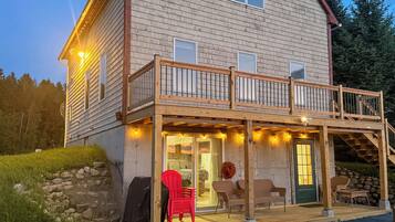 A two-story wooden house with a covered porch, outdoor seating, and a stone wall.