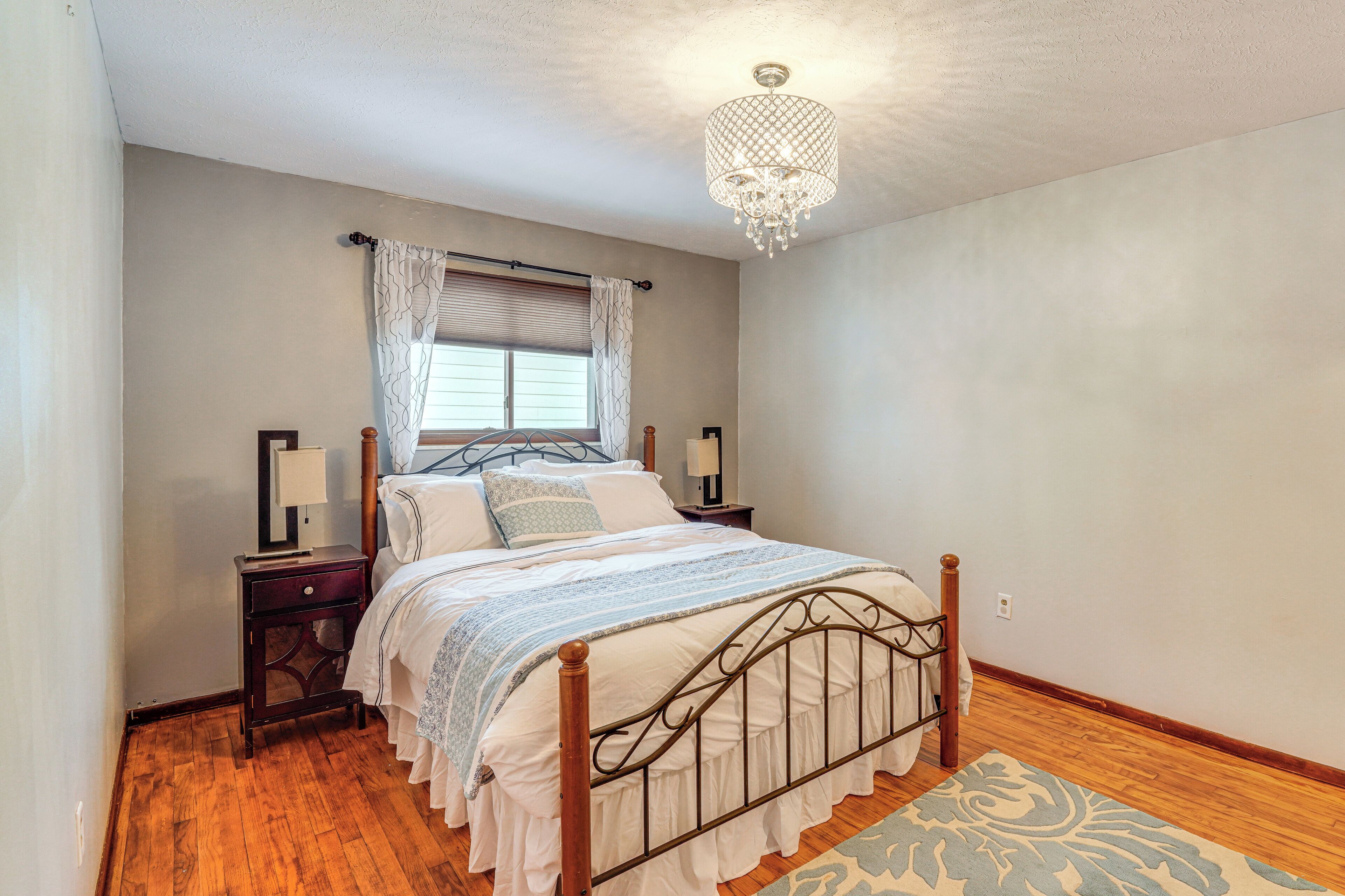 Un dormitorio con una cama de madera, un candelabro, una ventana con cortinas y una mesita de noche con una lámpara.