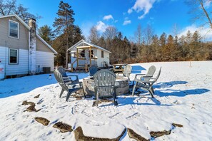 A snow-covered outdoor seating area with Adirondack chairs and a fire pit, surrounded by a house and trees. - Year-round Fun on Lake Sheridan! Home w/ Patio (Nicholson)