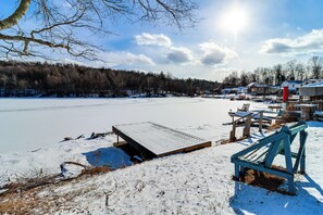 A snow-covered dock extending into a frozen lake, surrounded by bare trees and a few buildings in the background.