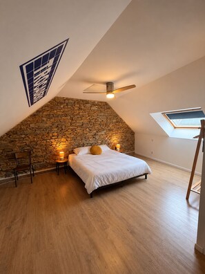 A bedroom with a sloped ceiling, a stone wall, a bed with white bedding, a ceiling fan, and a skylight. - Paris Cottage – Elegant stay & pool access – Les Gîtes du Petit Marcel (Soudan)