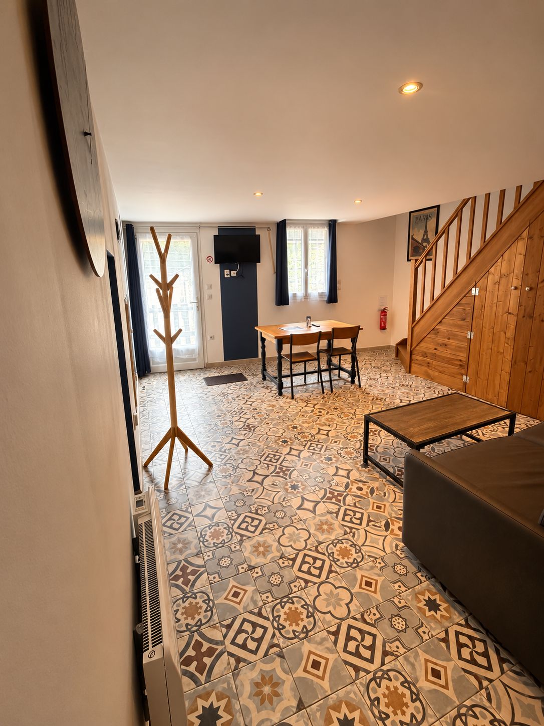 A room with a patterned floor, a dining table with chairs, a coat rack, a staircase, and a television mounted on the wall.