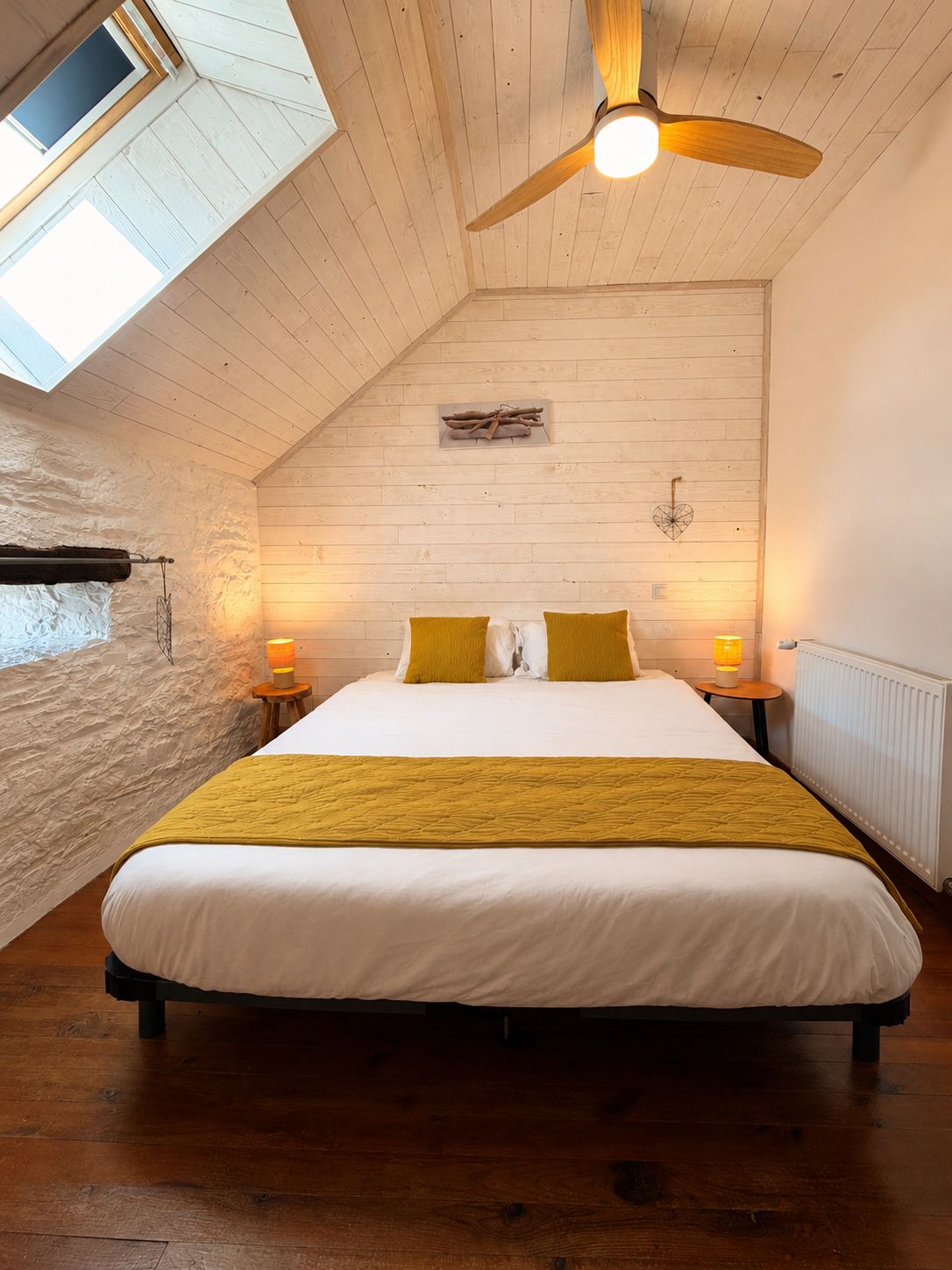 Quarto com teto inclinado, cama com roupa de cama branca e amarela, duas luminárias de cabeceira, piso de madeira e janela com vista.
