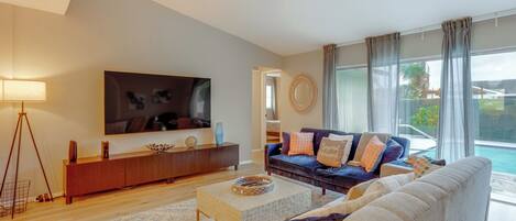 A modern living room with a large flat-screen TV, a dark blue sofa with patterned cushions, a beige sofa, a wooden coffee table, and a large window with a view of a swimming pool.