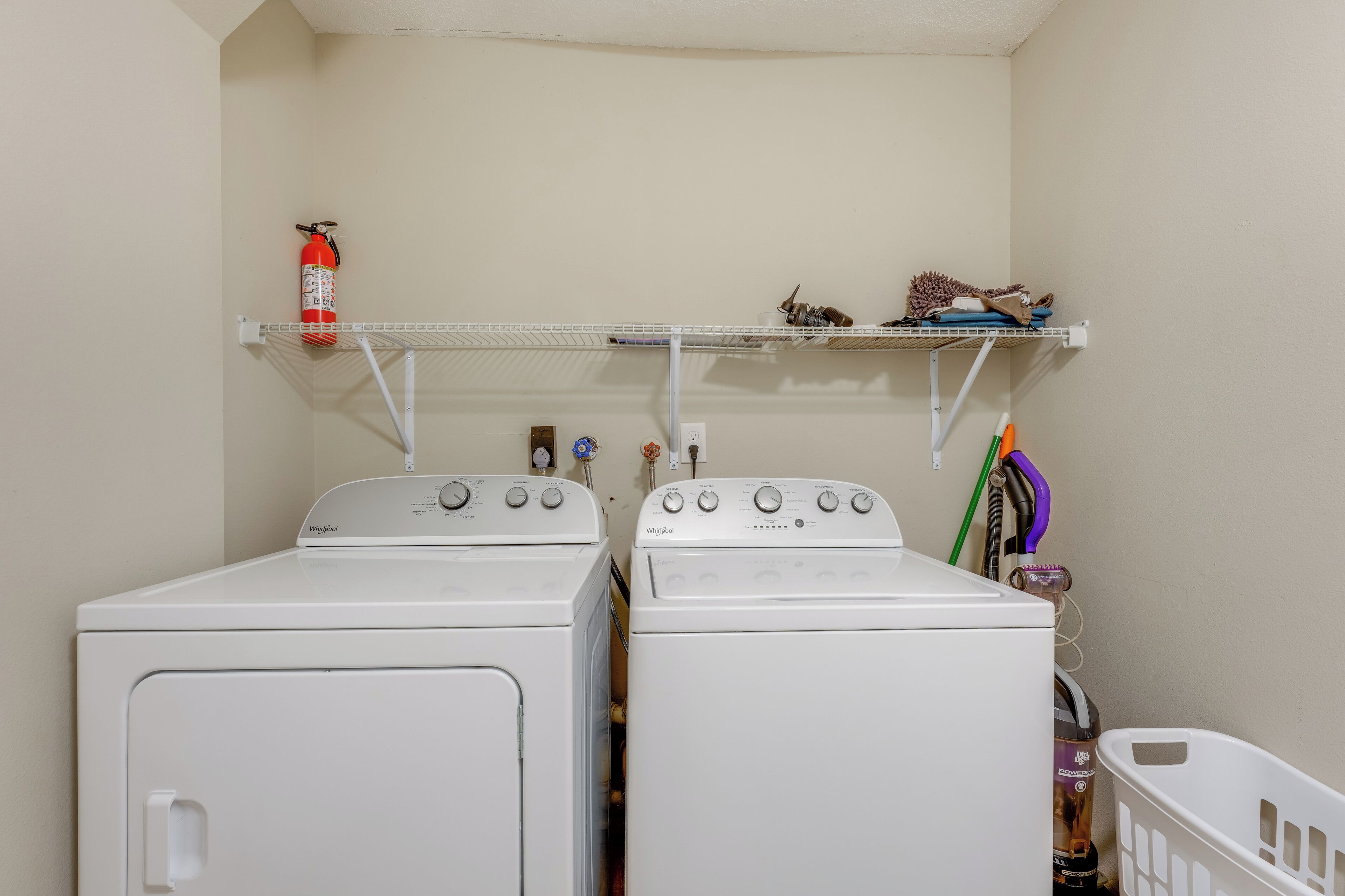 Two white Whirlpool front-loading washing machines in a laundry room with a shelf above holding a fire extinguisher and a small stuffed animal.
