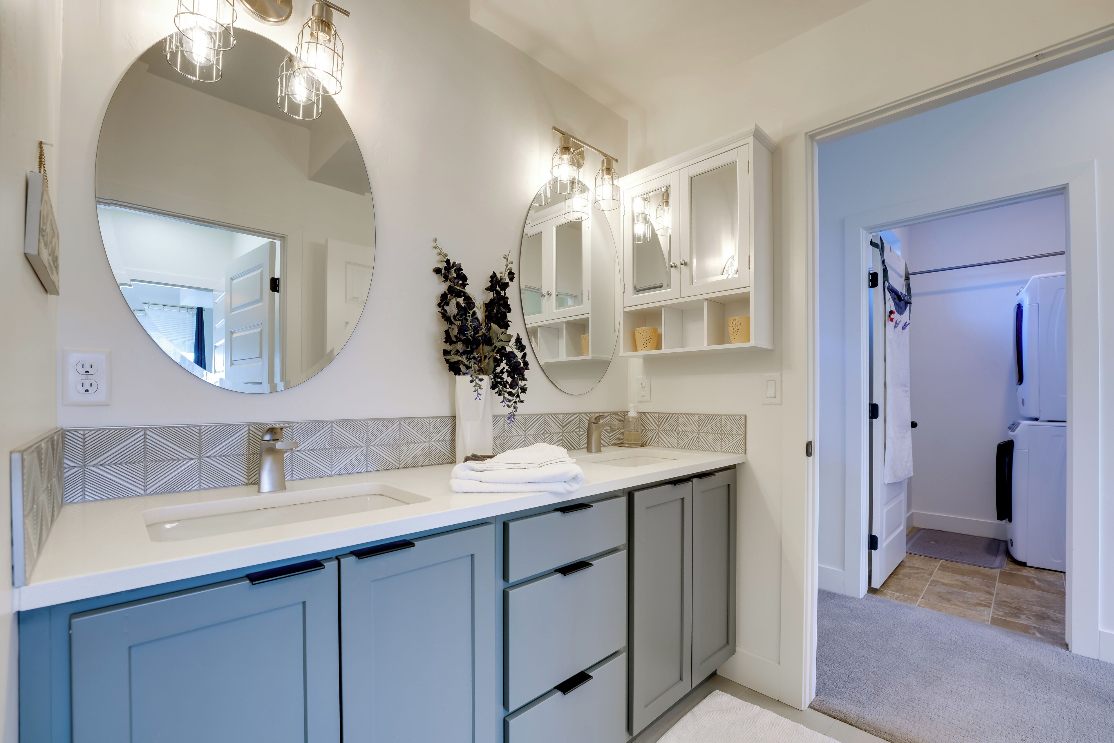 A modern bathroom with a large round mirror, two pendant lights, a white sink, and grey cabinets.