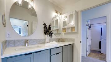 A modern bathroom with a large round mirror, two pendant lights, a white sink, and grey cabinets.