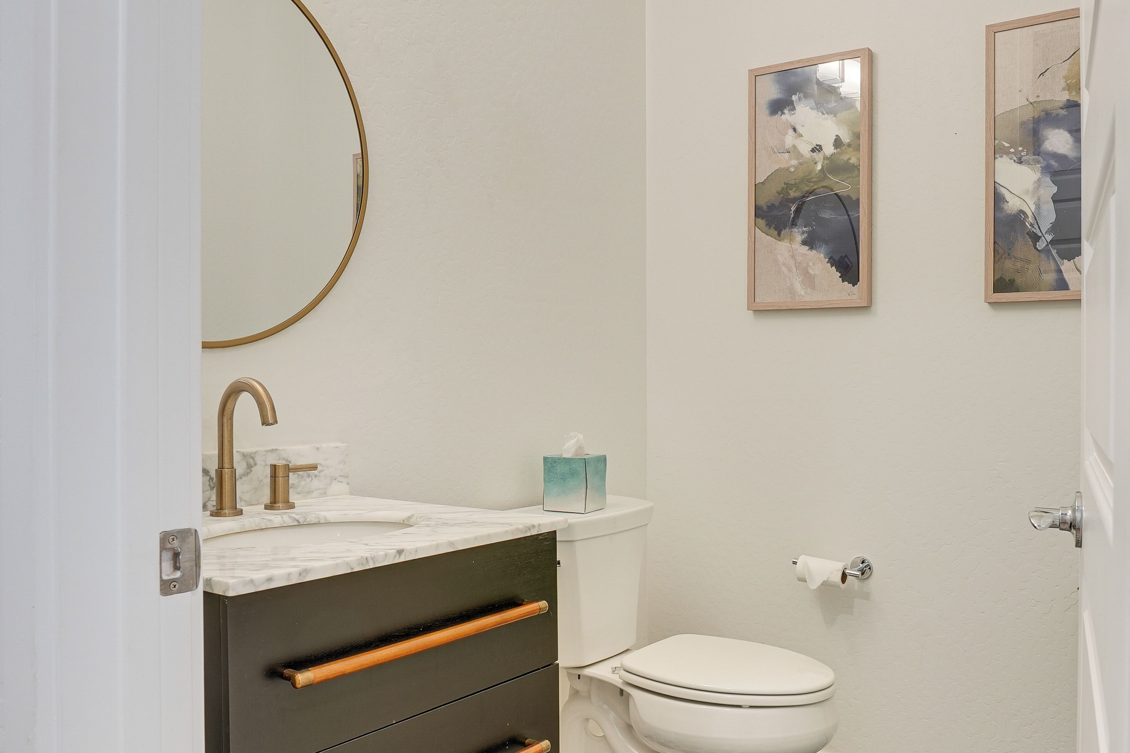 Un bagno con un water bianco, un rubinetto dorato, un lavabo in marmo e un cassetto in legno. Alle pareti, due quadri astratti incorniciati.