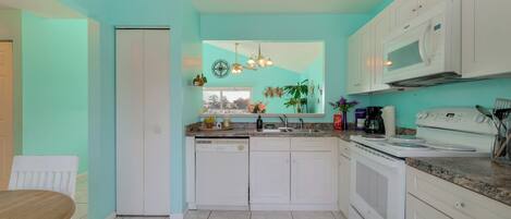 Cocina con alacenas blancas, microondas y un lavamanos. Se ve una ventana con plantas y una lámpara colgante.