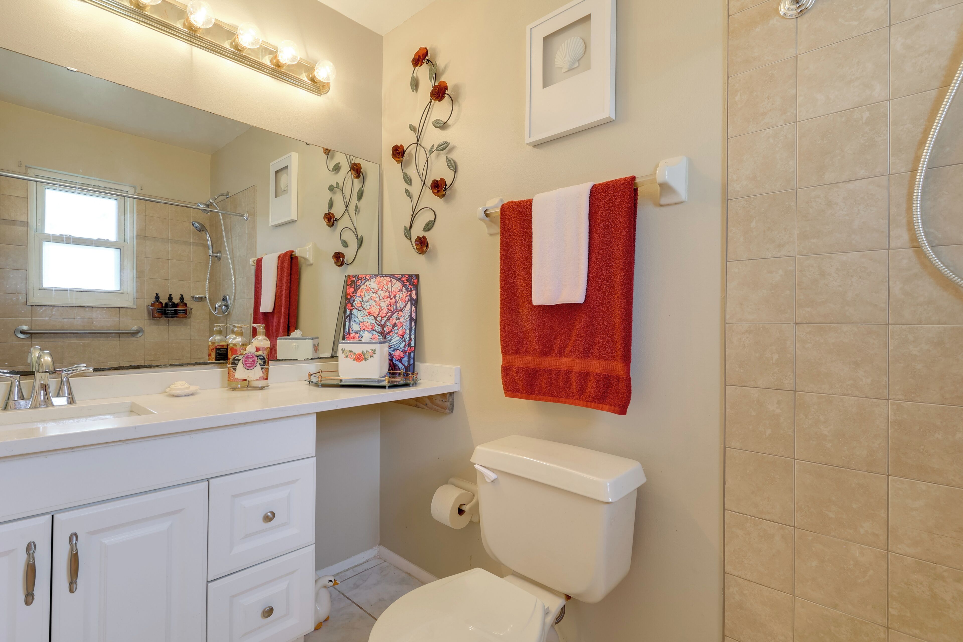 Une salle de bain avec un lavabo blanc, des toilettes, une douche et un miroir. Des serviettes rouges sont accrochées au mur.