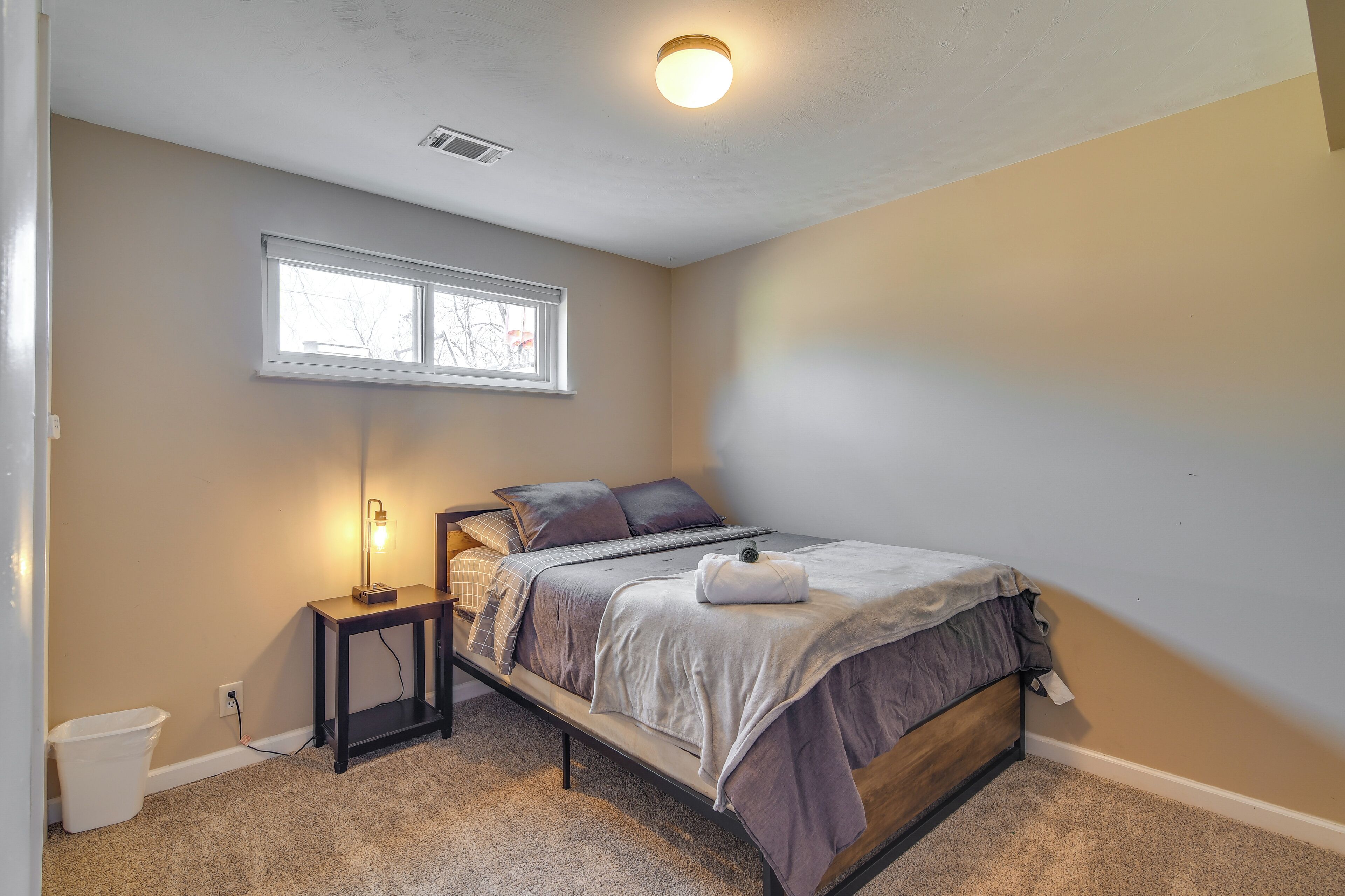 A bedroom with a single bed, a bedside table, a lamp, a window, and a carpeted floor.